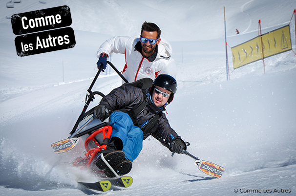 Deux hommes souriants descendent à toute allure une piste de ski. L'un d'entre eux est est en tandem-ski.