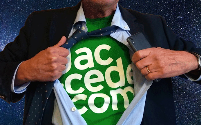 Gros plan sur le buste d'un homme (vu de face) portant un costume cravate et tenant un téléphone dans sa main. Il ouvre sa chemise qui laisse apparaître son t shirt Jaccede 